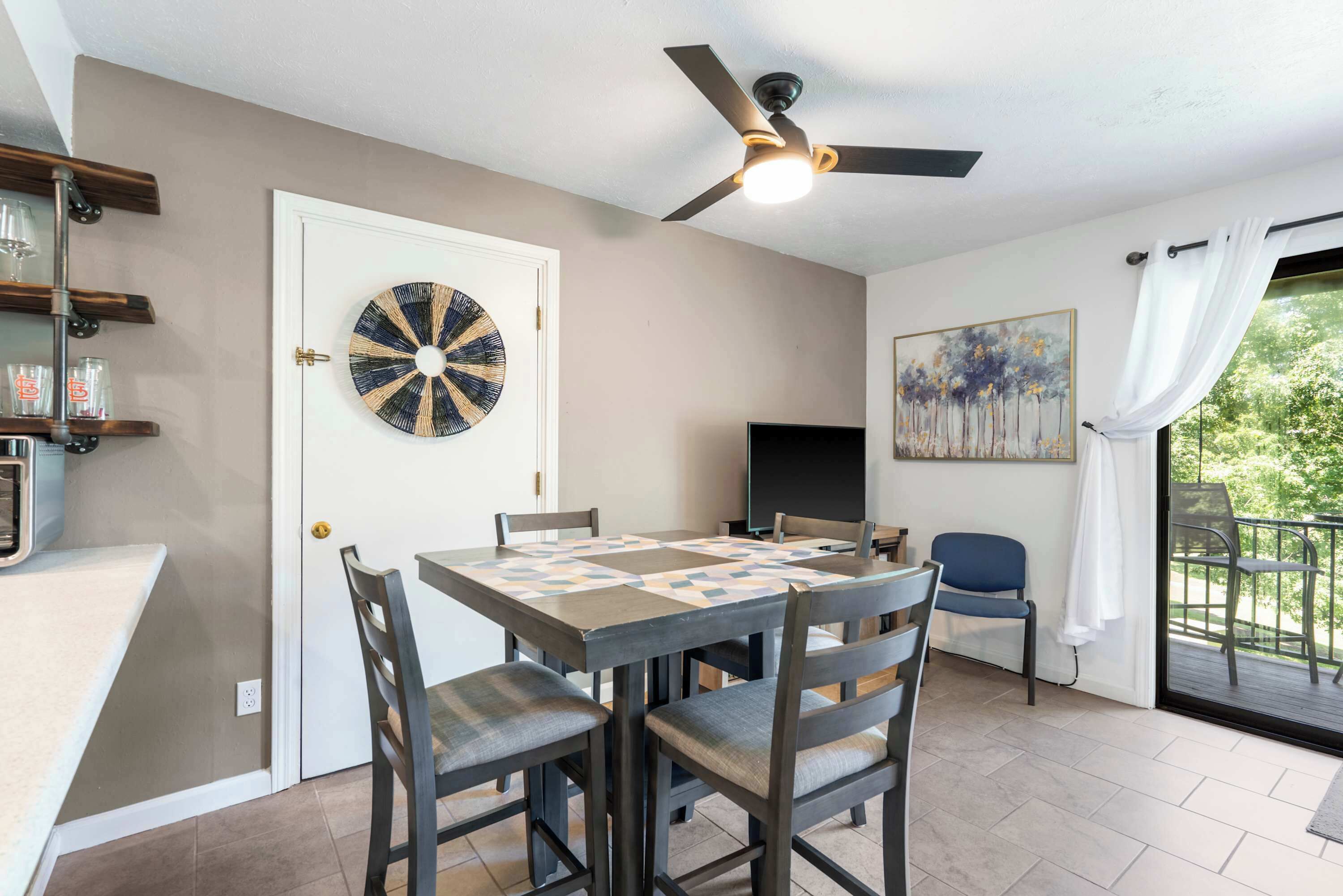 A dining area with a ceiling fan, a wall-mounted decorative plate, a painting, and a view of greenery through a window.