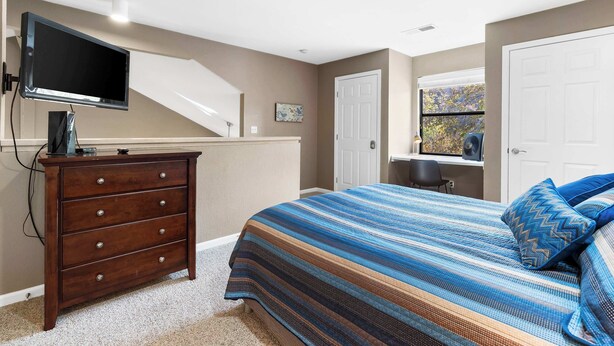 A bedroom with a bed, a dresser, a TV mounted on the wall, and a window with a view of trees. - Whispering Water @ Pointe Royale (Branson)