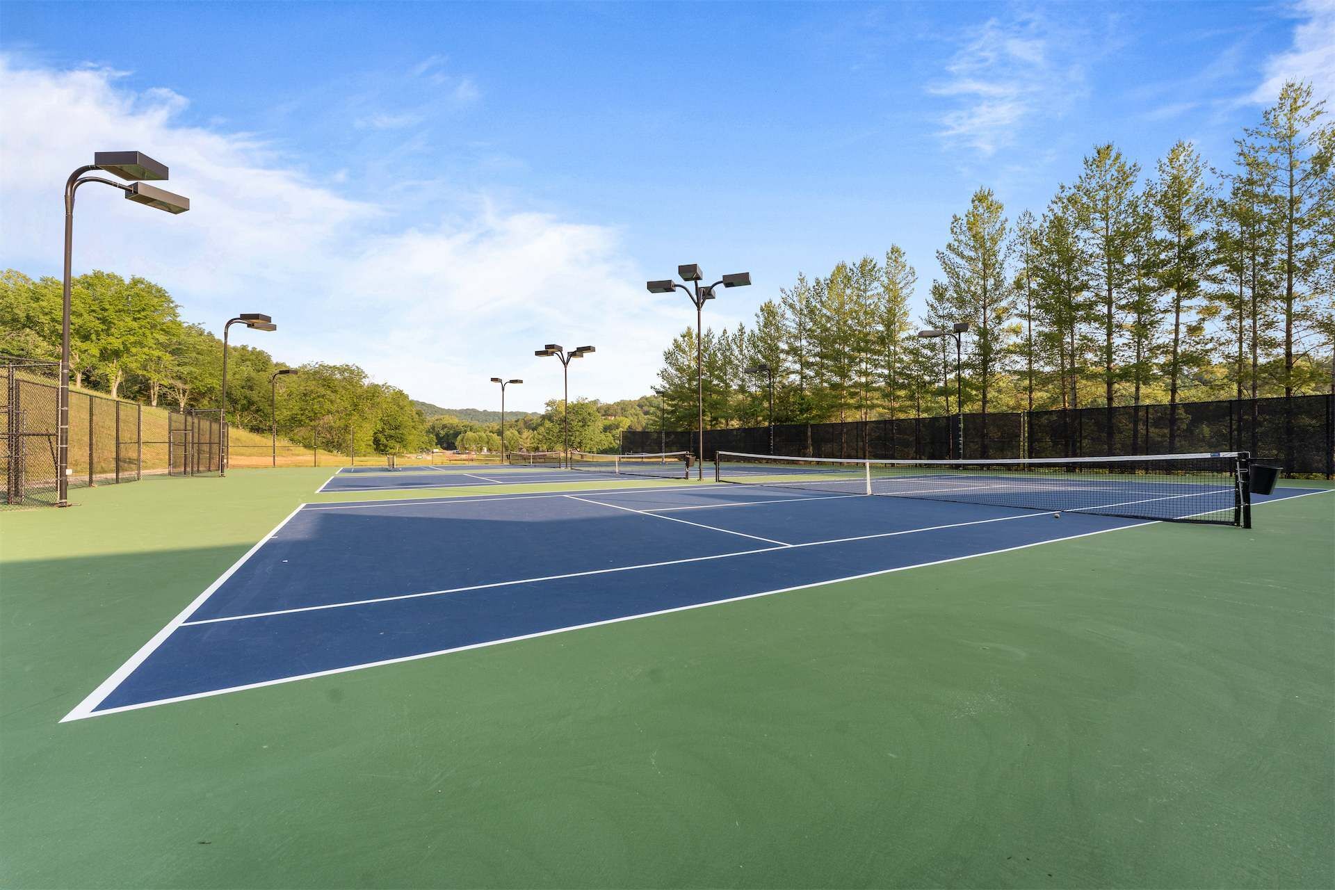 Un court de tennis extérieur, doté de plusieurs mâts d’éclairage, entouré d’arbres et d’une clôture.