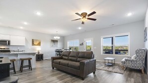 A spacious living area with a brown leather sofa, a dining table with stools, a kitchen area, and a large window offering a view of the outdoors. - Stonebridge Escape Sleeps 8! (Branson West)
