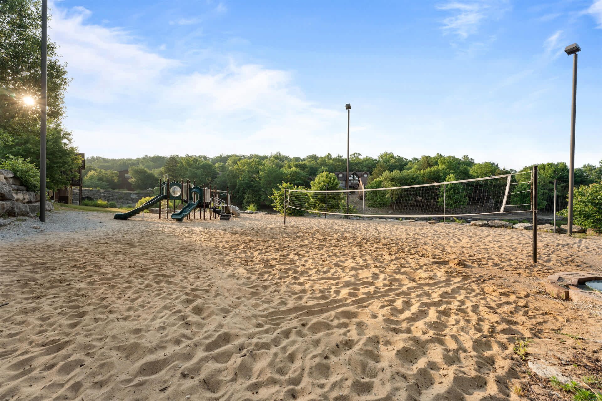 Una playa arenosa con una red de voleibol, un parque infantil con toboganes y árboles al fondo.