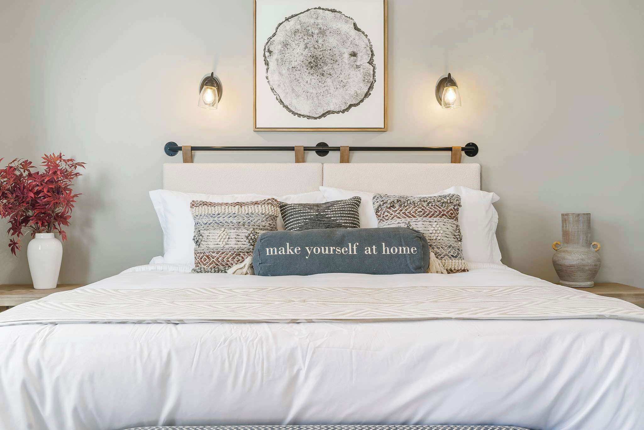 A neatly made bed with a white comforter and multiple pillows, including one with a motivational quote. There is a framed artwork above the bed and two wall-mounted lights on either side.