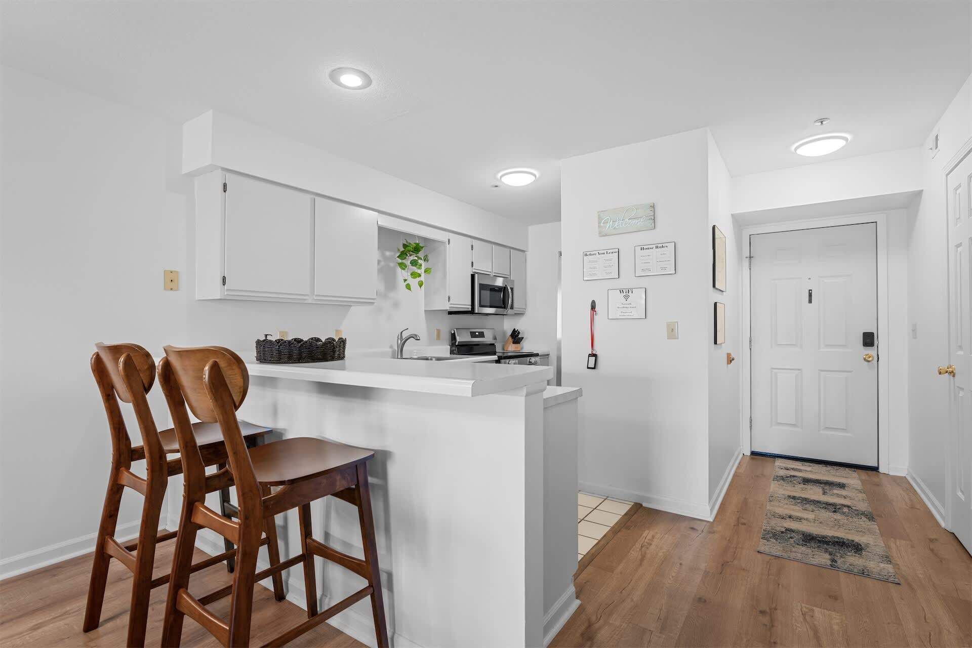 A modern kitchen with white cabinetry, a built-in microwave, and a bar area with two wooden bar stools.
