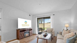 A modern living room with a flat-screen TV, a fireplace, a sofa, a coffee table, and a view through large glass doors. - Royal Ridge Modern Condo for 4 (Branson)