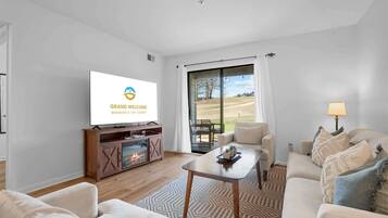 A modern living room with a flat-screen TV, a fireplace, a sofa, a coffee table, and a view through large glass doors.