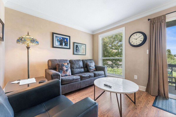 A living room with a sofa, a coffee table, and a lamp. There are framed pictures on the wall and a clock. - Pointe Royale Getaway - Fore 4 (Branson)