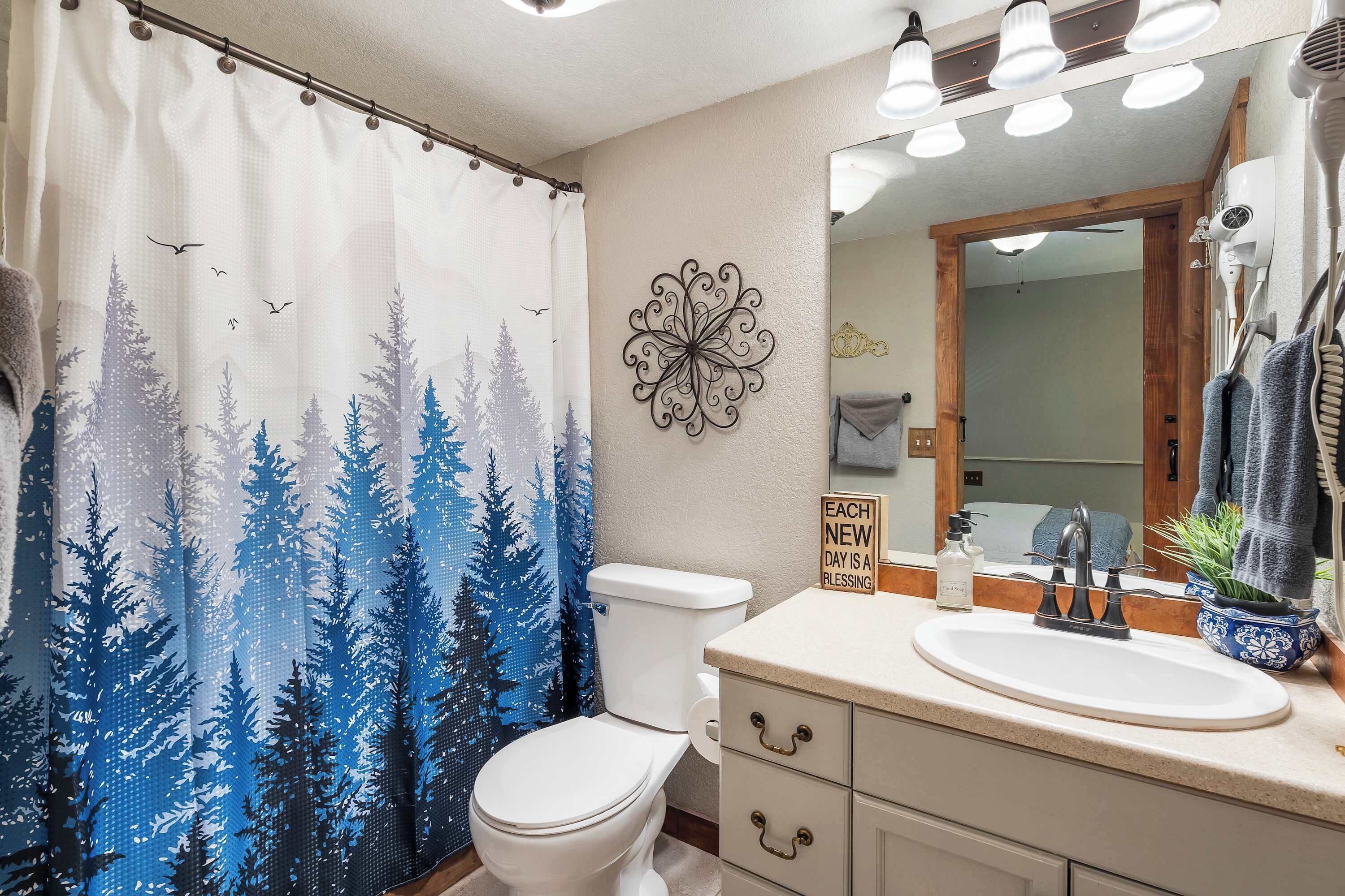 A bathroom with a white toilet, a shower curtain with a forest pattern, a wall-mounted decorative piece, a mirror with a wooden frame, and a sink with a soap dispenser.