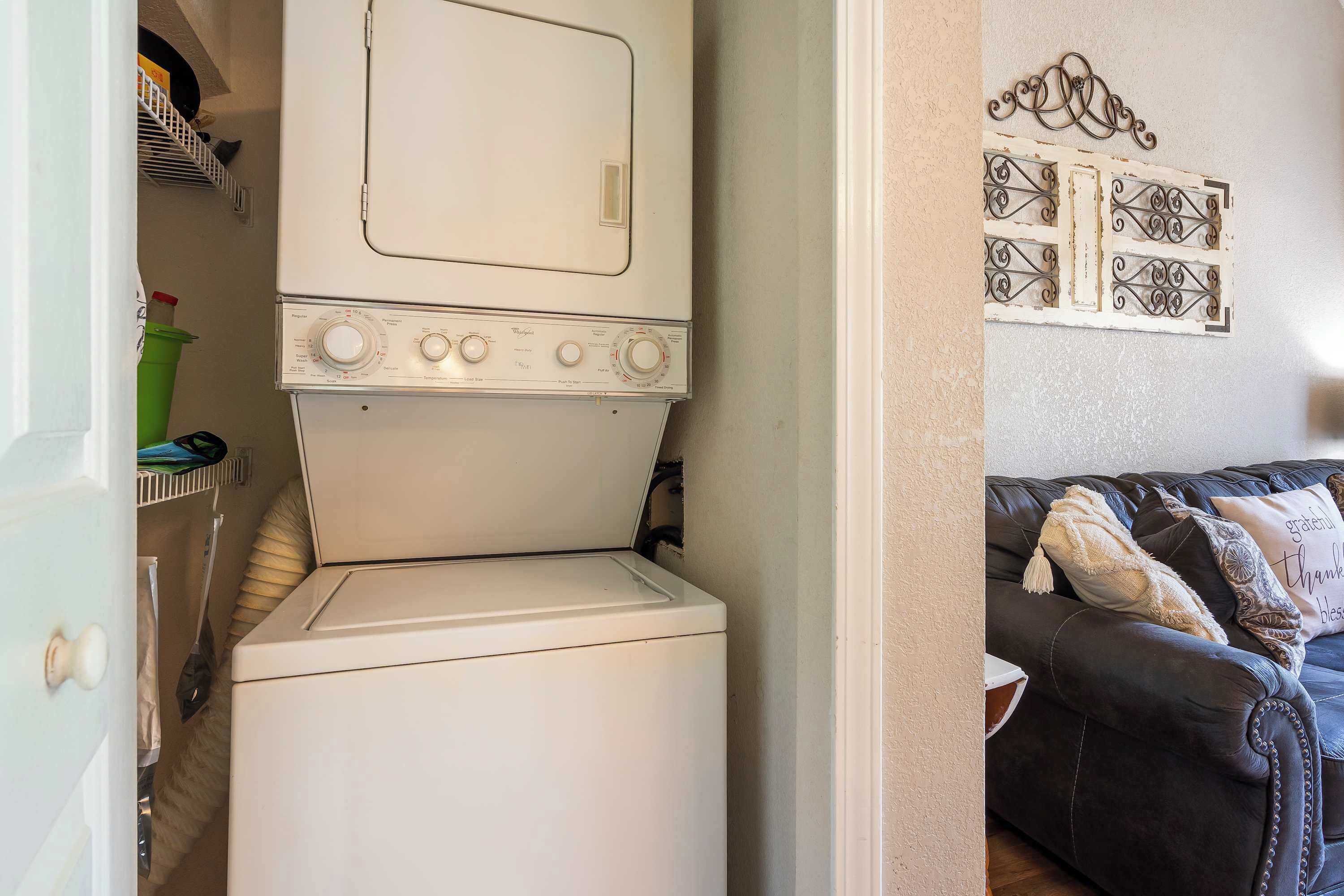Un espacio pequeño para lavandería con lavadora y secadora de carga frontal, una cesta de ropa verde y un sofá de cuero negro con almohadones decorativos.