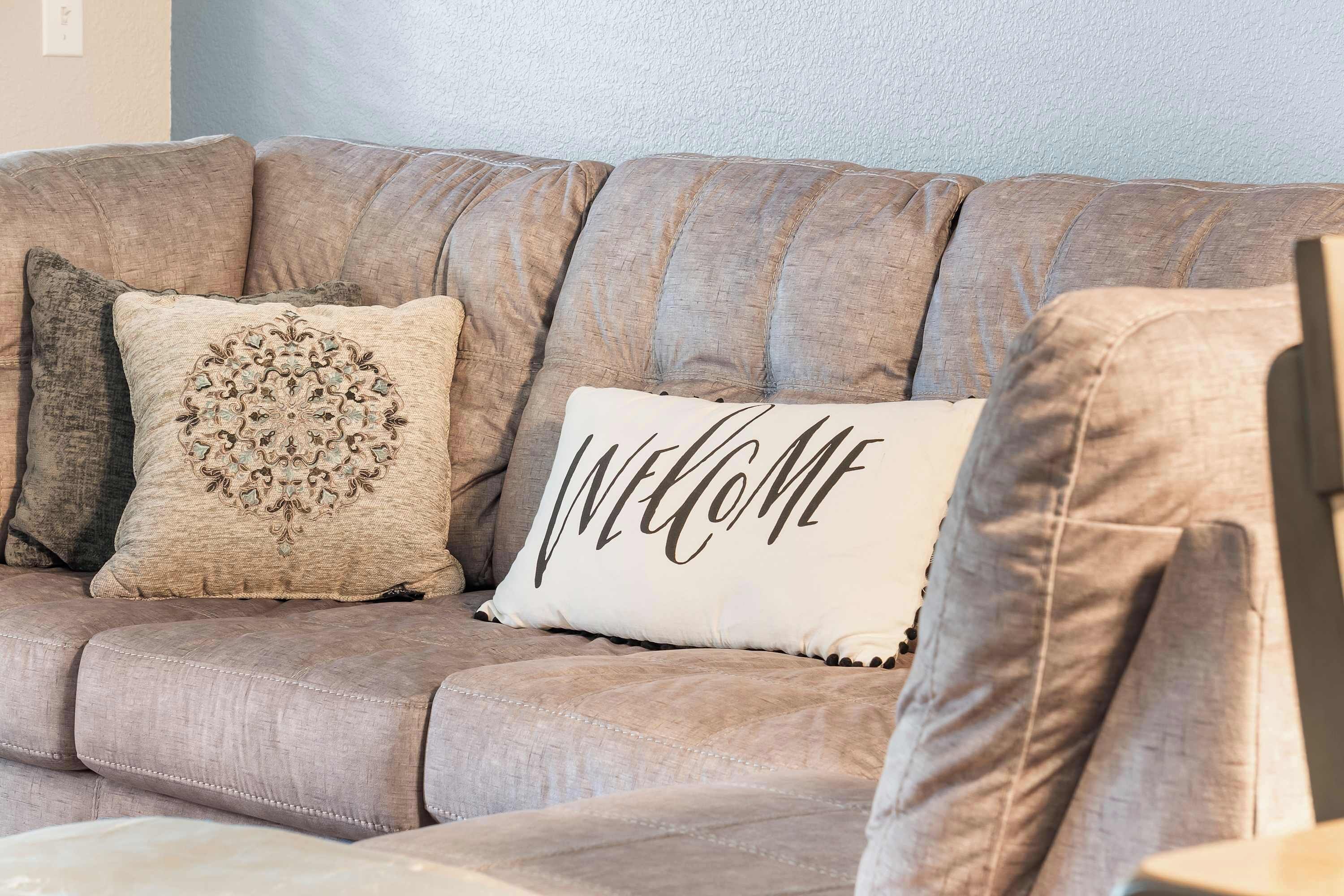 Een beige bank met een 'WELCOME' kussen en een decoratief kussen met een cirkelpatroon.