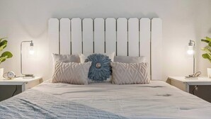 Une chambre à coucher avec une tête de lit blanche, un grand lit recouvert d’une couette bleue et blanche, deux tables de chevet avec des lampes, et une fleur bleue décorative posée sur le lit.
