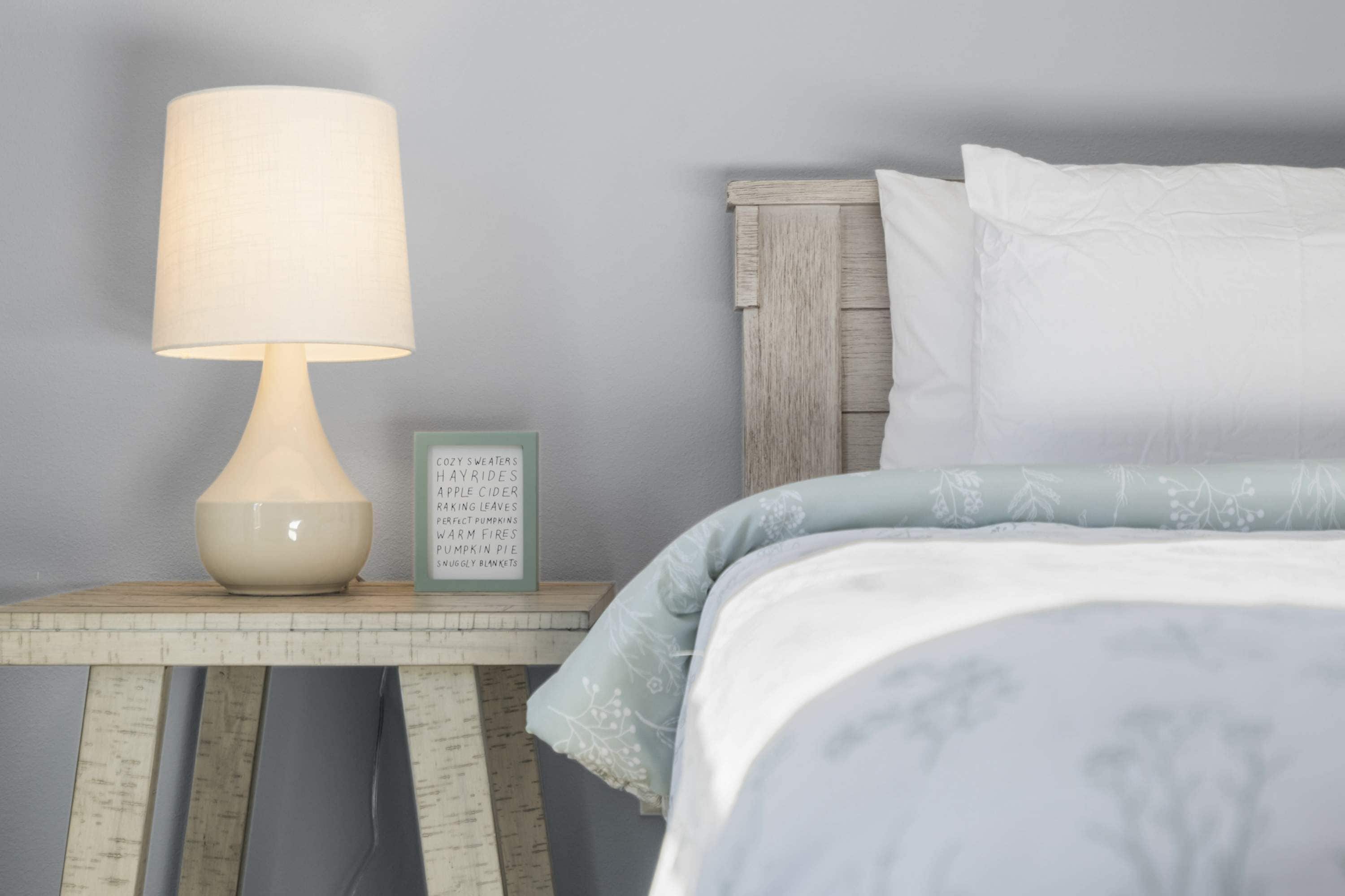 Un lit bien fait, recouvert d’une couvre-lit bleu clair et agrémenté d’oreillers blancs, une table de chevet avec une lampe et un panneau indiquant les articles de saison.