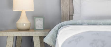 Un lit bien fait, recouvert d’une couvre-lit bleu clair et agrémenté d’oreillers blancs, une table de chevet avec une lampe et un panneau indiquant les articles de saison.