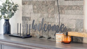 Zona de recepción con pared de piedra, un cartel que dice 'Welcome', una computadora portátil, una vela y un florero con hojas verdes.