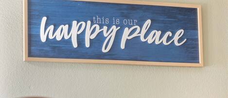 A framed sign with the text "this is our happy place" on a textured wall.