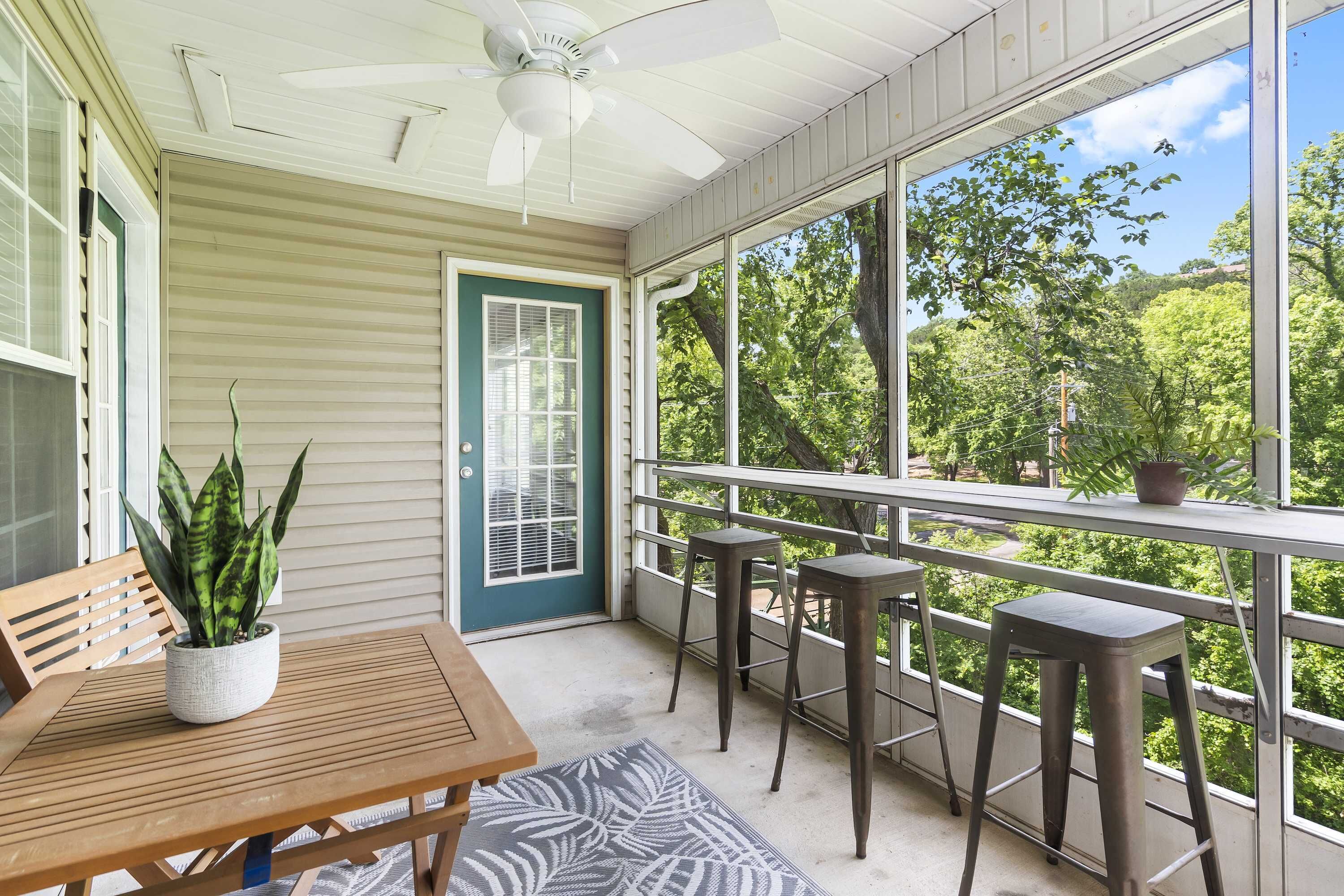 A covered porch with a table and two bar stools, a potted plant, and a view of trees and hills.