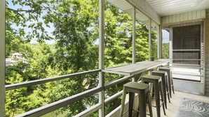 A balcony with a view of a forested area, a table, and bar stools. - Lakeside Bliss Retreat 2 BDR (Branson)