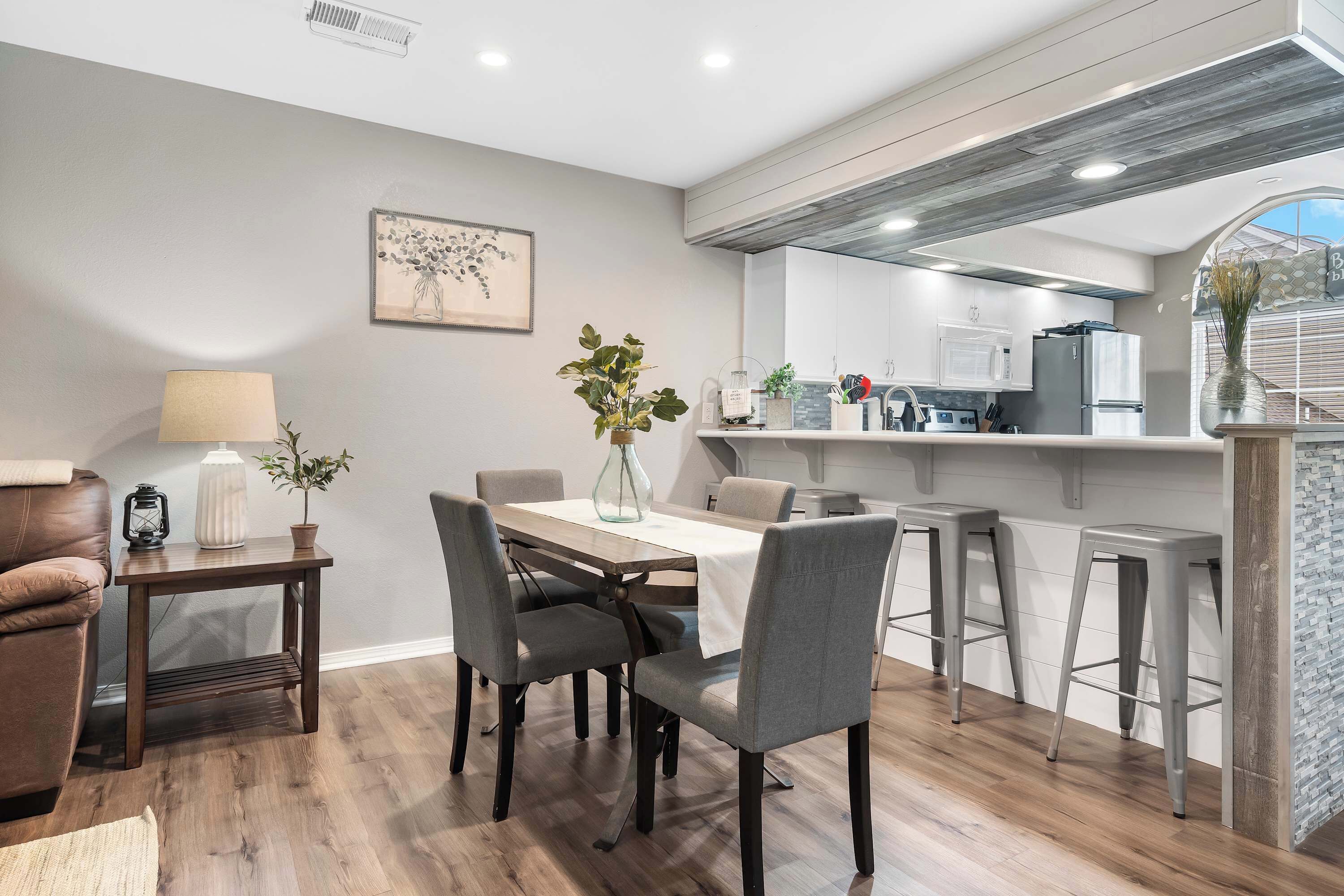A modern kitchen with white cabinets, a dining area with a wooden table and grey chairs, and a small side table with a lamp and a plant.