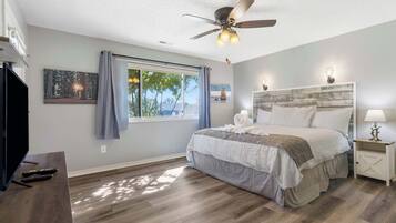 A bedroom with a bed, a ceiling fan, a window with curtains, a TV, and a wall-mounted artwork.