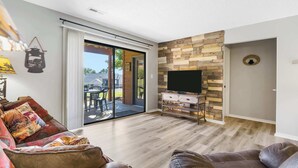 A modern living room with a sofa, a TV on a wooden cabinet, and a sliding glass door leading to an outdoor patio.