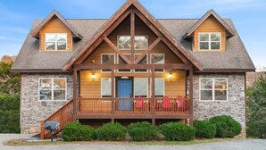 A two-story log cabin with a stone foundation, a wooden front porch with red chairs, and a stone chimney. - Kemper's Cabin in the Ozarks - Onsite Amenities (Branson)