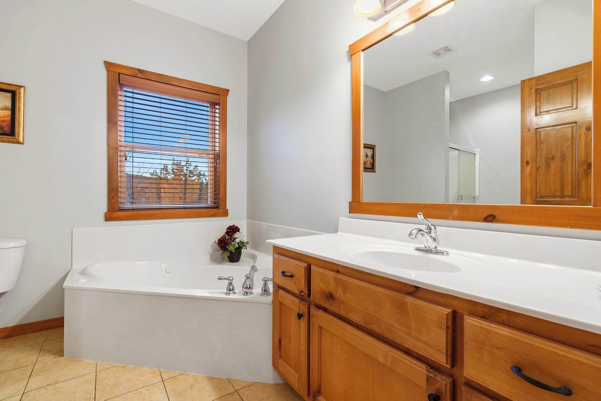 A bathroom with a bathtub, two sinks, wooden cabinets, a large mirror, and a window with blinds.