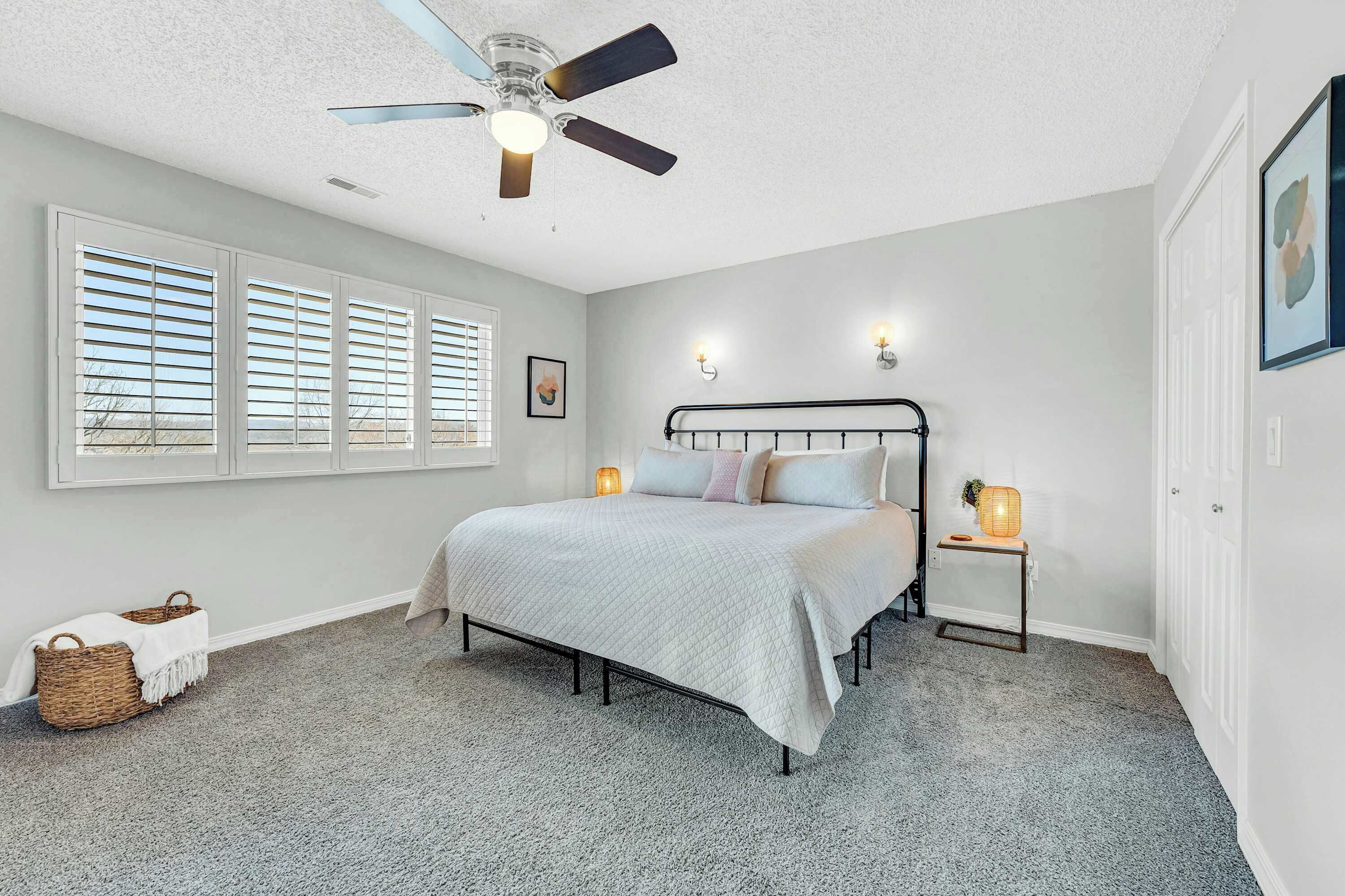 A bedroom with a bed, a ceiling fan, a window with blinds, a bedside table with a lamp, and a wall-mounted light fixture.
