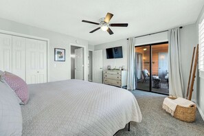 Ein Schlafzimmer mit einem Bett, einem Deckenventilator, einem Schminktisch, einem Fernseher und einer Glastür, die auf einen Balkon führt.