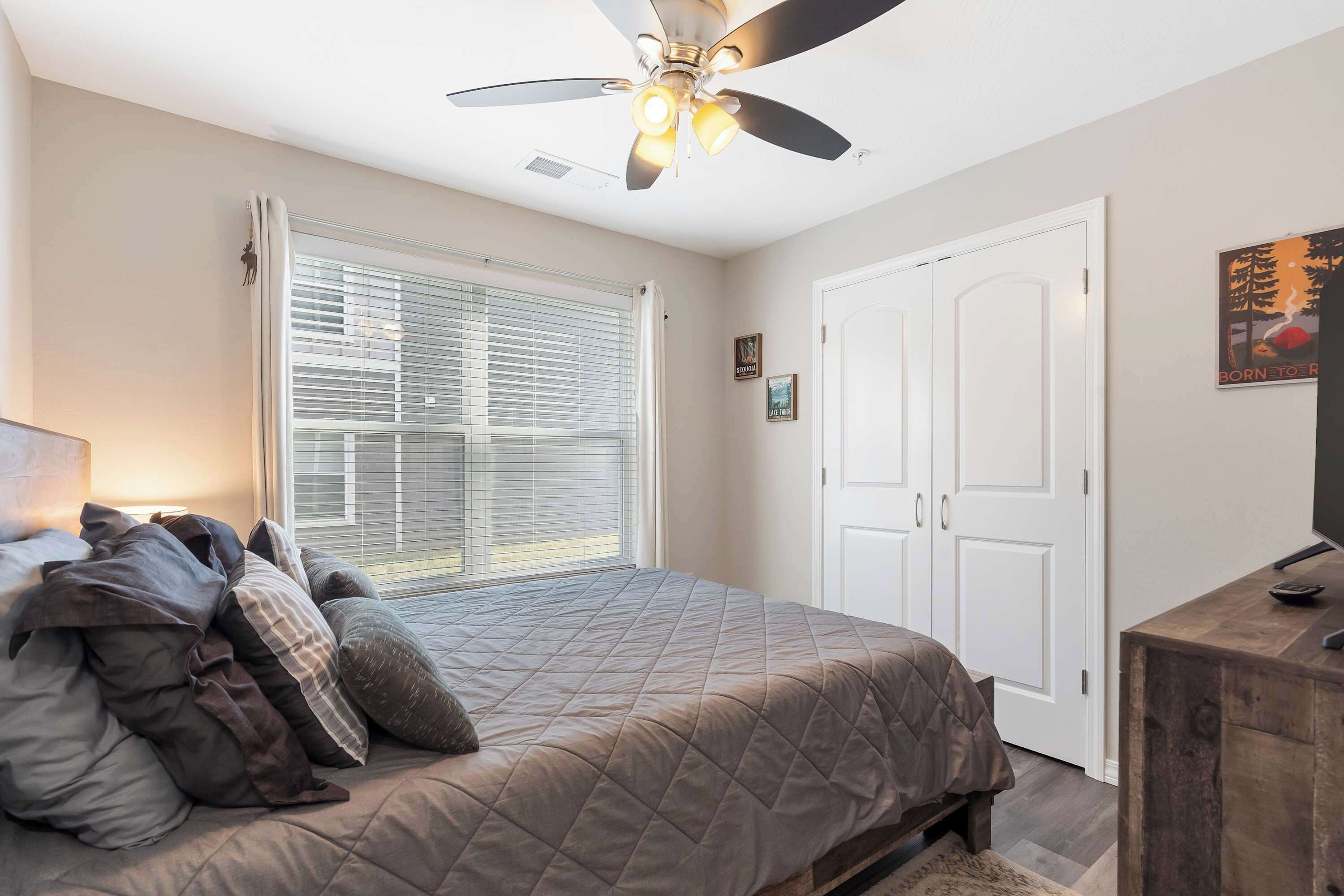 A bedroom with a bed, a ceiling fan, a window with blinds, a door, and a dresser.