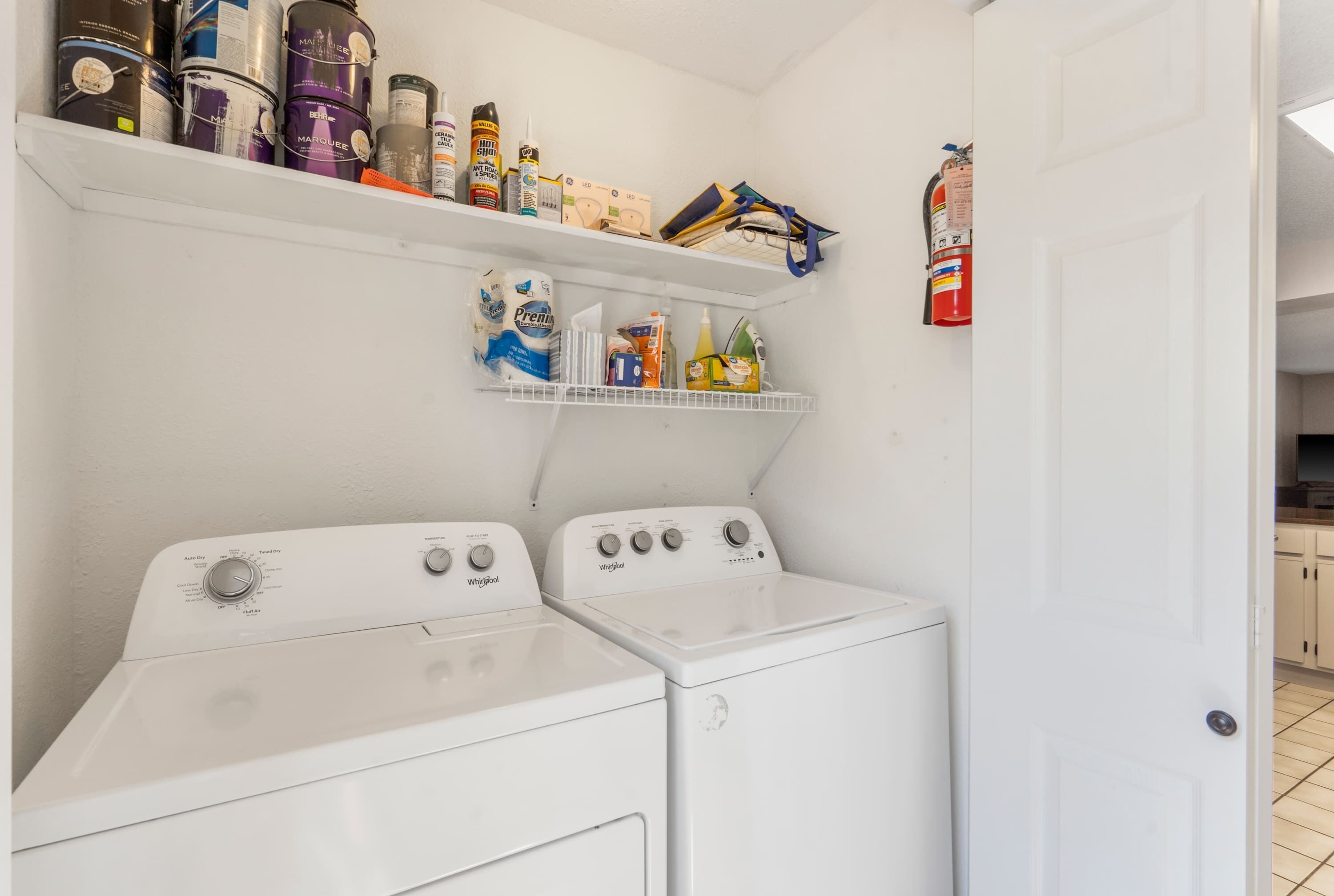 A laundry room with two white Whirlpool washers and dryers, shelves above containing cleaning supplies, and a fire extinguisher mounted on the wall.