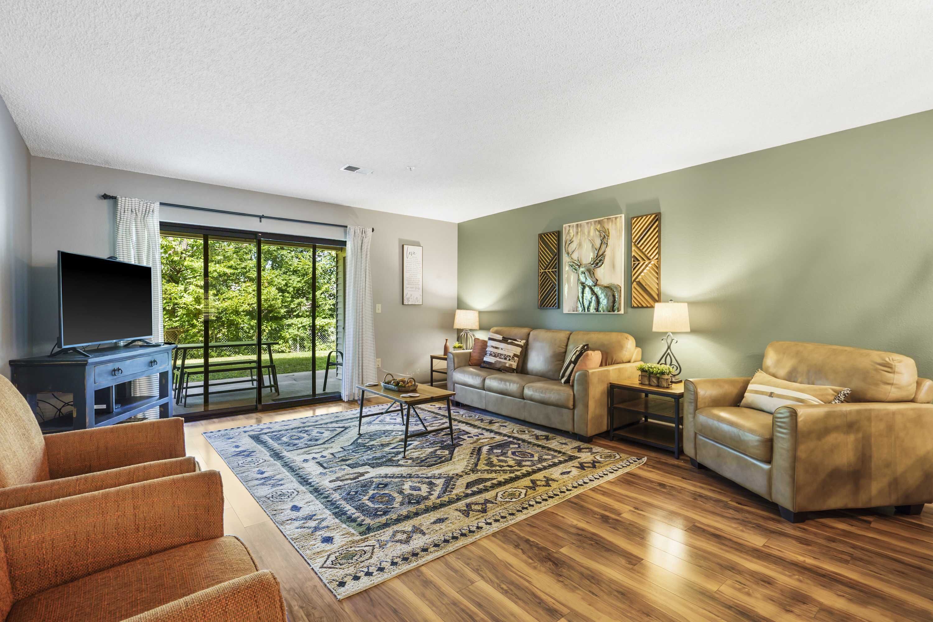 A spacious living room with a large window, leather sofas, a flat-screen TV, and a patterned rug.