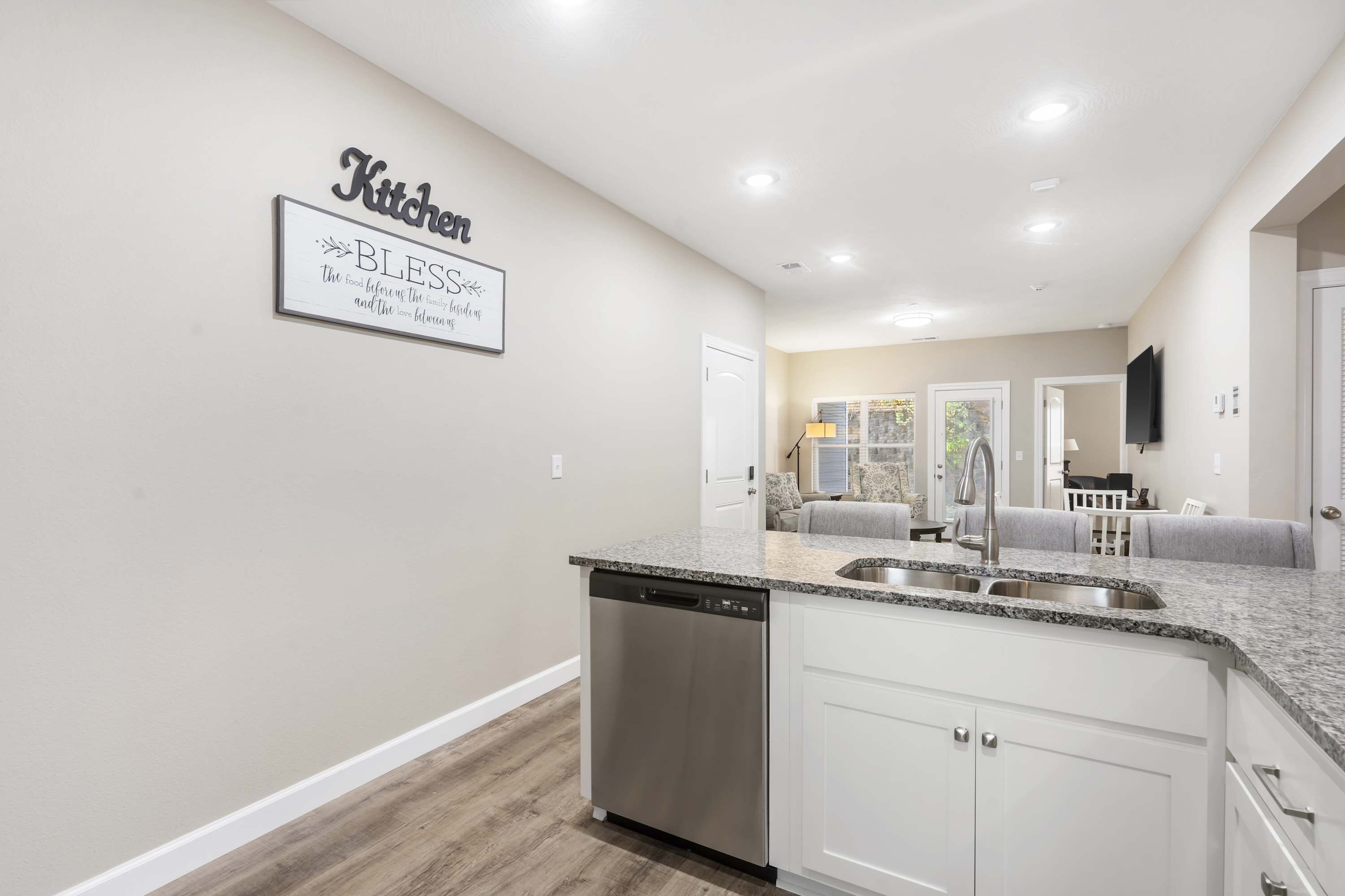 A modern kitchen with a granite countertop, stainless steel sink, and a dishwasher. There is a sign on the wall that reads "Kitchen" and "Blessed."