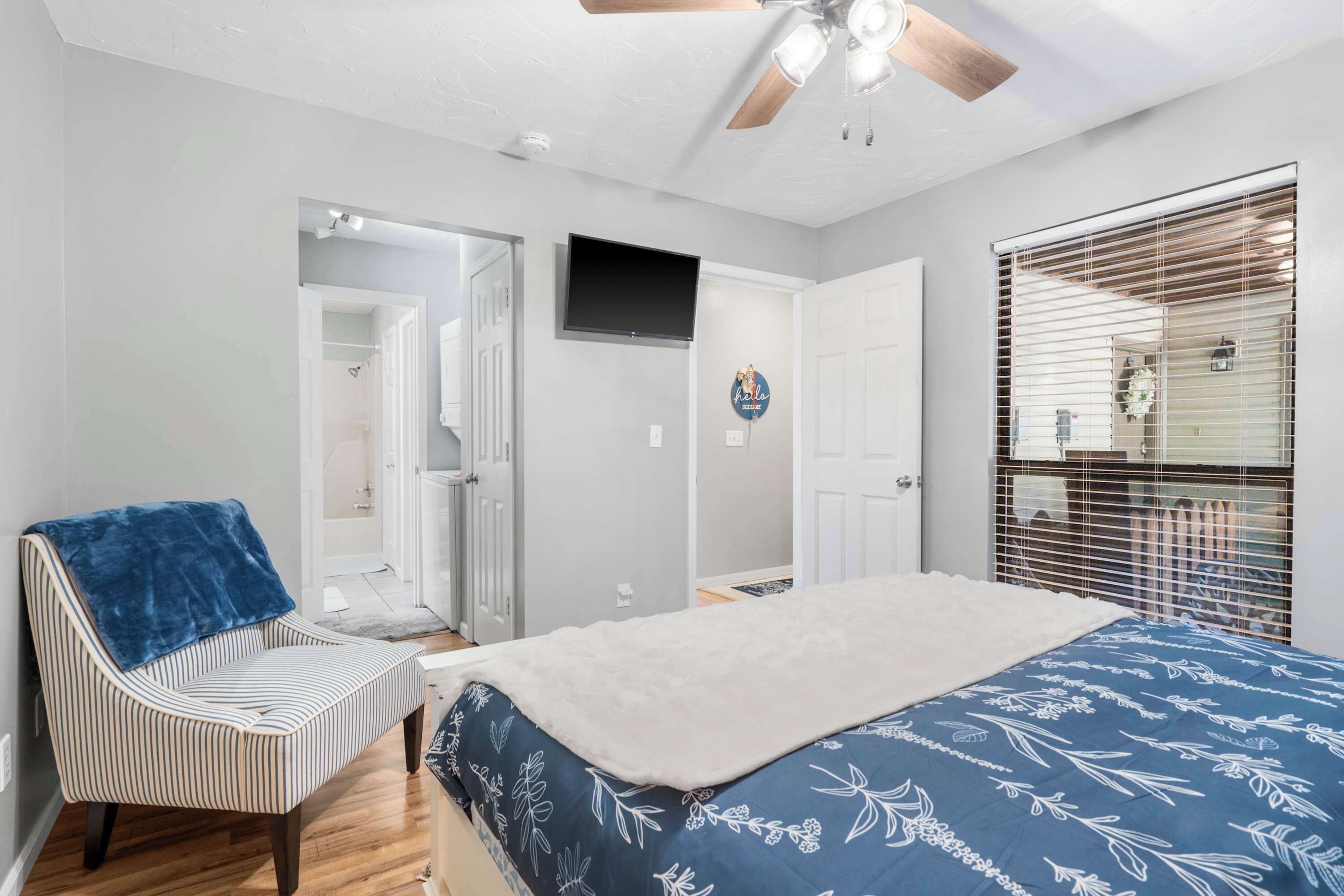 A bedroom with a bed, a chair, a ceiling fan, a TV, and a window with blinds.