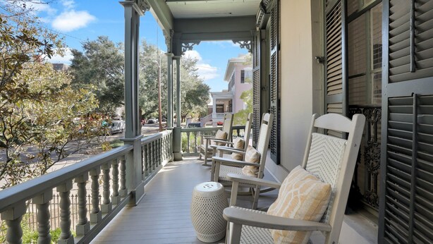A porch with wicker chairs and a round ottoman, overlooking a street with trees and buildings.