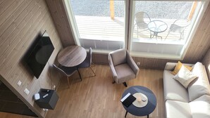 A modern living room with a TV, two round tables, a sofa, and a large window overlooking a deck. - Åtte Årstider Lodge (South Varanger)