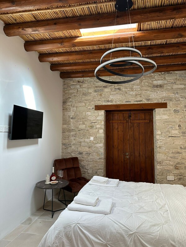 A bedroom with a stone wall, wooden ceiling, a bed with white bedding, a brown armchair, a small round table with a bottle and glass, and a TV mounted on the wall.