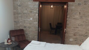 A bedroom with a stone wall, a bed with white linens, a brown chair, a small round table with a bottle and glasses, and a door leading to a dining area.