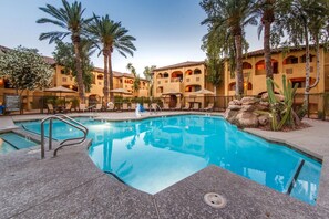 Relax by the serene pool surrounded by lounge chairs and umbrellas, amidst a Mediterranean-inspired setting with palm trees and cacti.