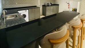 A modern kitchen with grey cabinets, a black countertop, a built-in washing machine, and a bar area with wooden stools. - Baithans Furnished Apartments (Ajman)