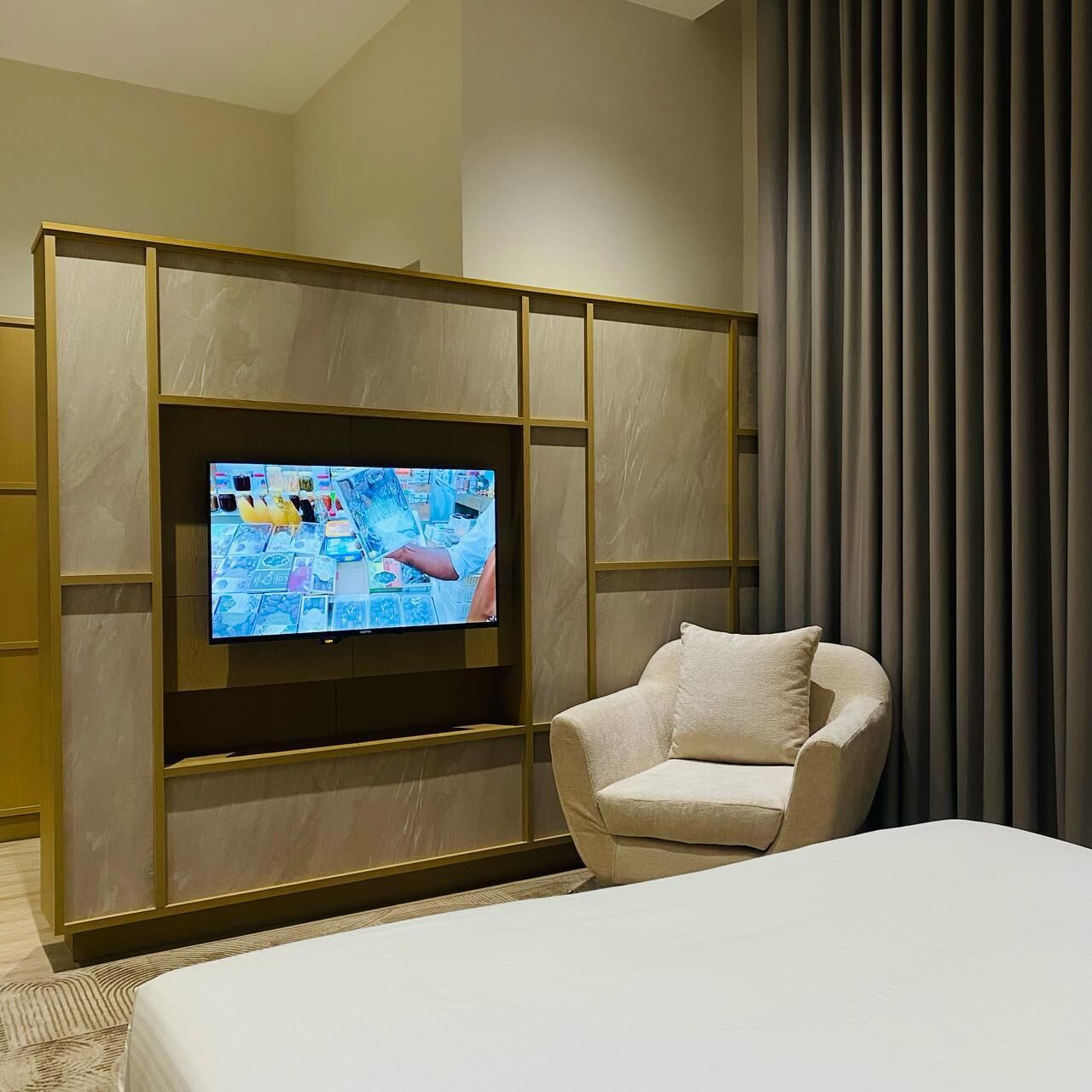 A hotel room with a large flat-screen TV mounted on a wooden panel wall, a beige armchair, and a large bed with a white comforter.