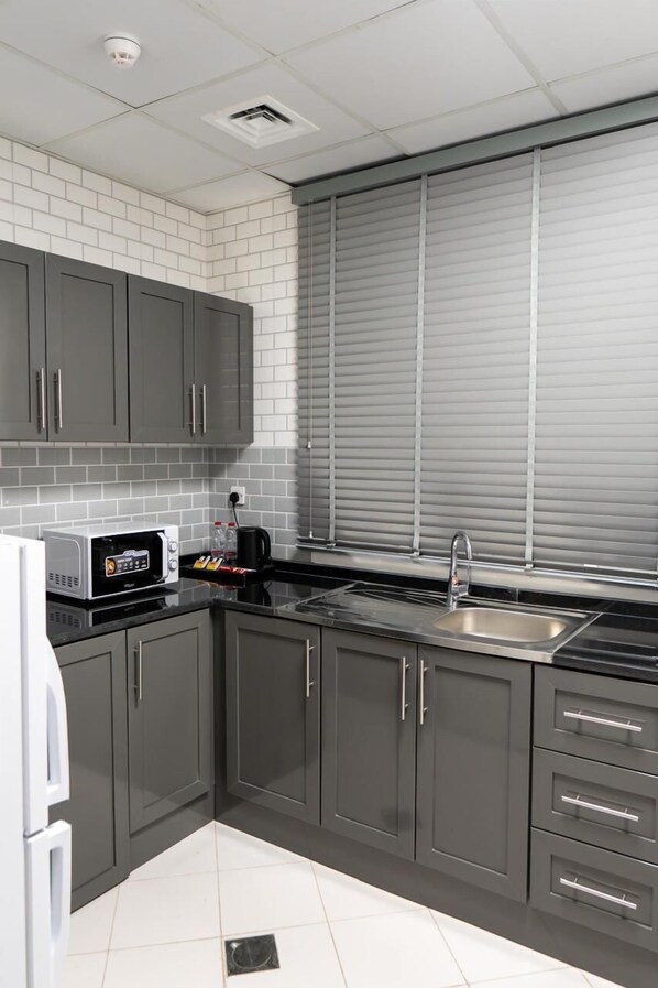 A modern kitchen with grey cabinets, a white tiled backsplash, a microwave, a sink, and a window with blinds. - Baithans Furnished Apartments (Ajman)