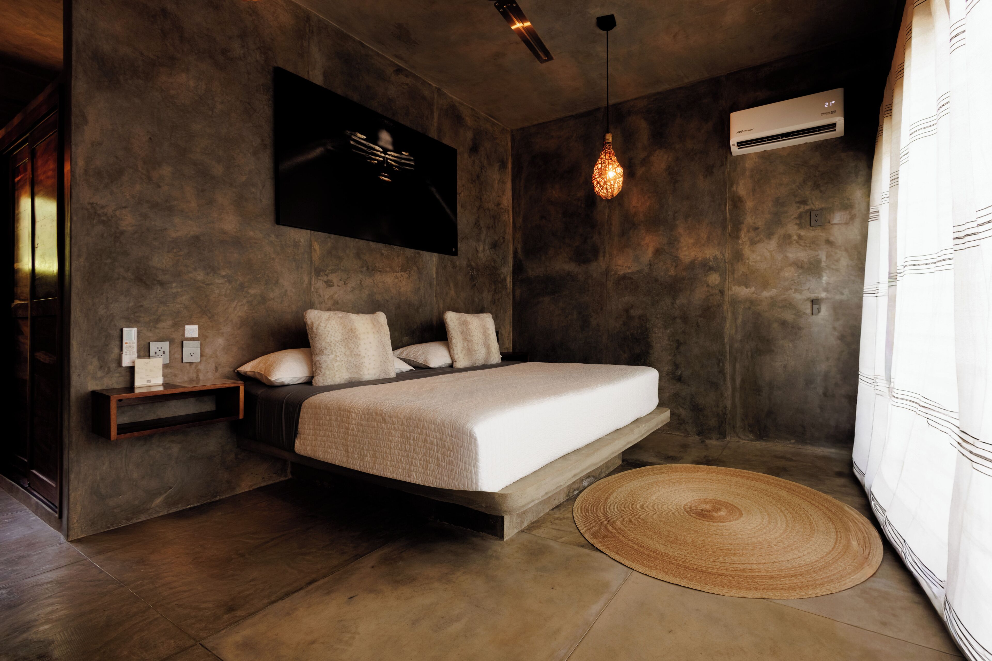 A modern bedroom with a bed, bedside tables, a wall-mounted air conditioner, and a hanging light fixture.