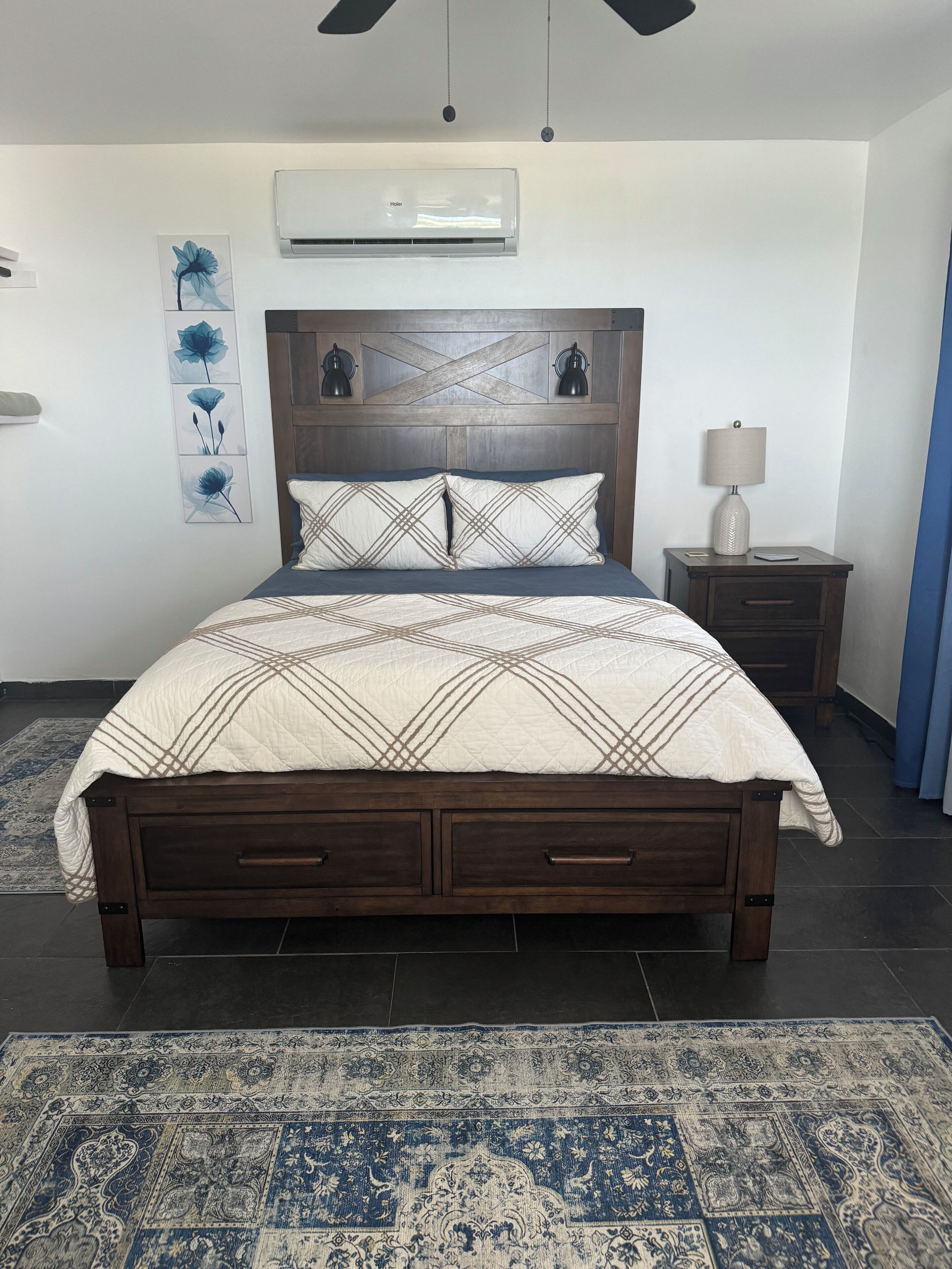 A bedroom with a wooden bed, a nightstand with a lamp, a wall-mounted air conditioner, and a decorative wall piece.
