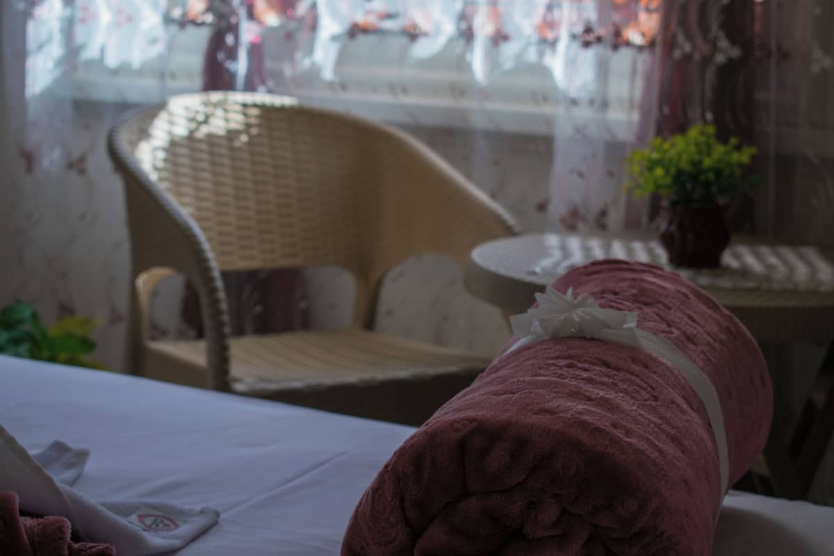 A bed with a white sheet and a pink towel rolled up at the foot. There is a wicker chair with a cushion in the background.