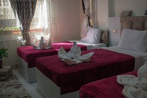 Ein Hotelzimmer mit zwei Betten, jeweils mit roter Bettdecke und weißen Kissen. Es gibt ein Fenster mit Vorhängen, einen Nachttisch mit Pflanze und eine an der Wand montierte Lampe.