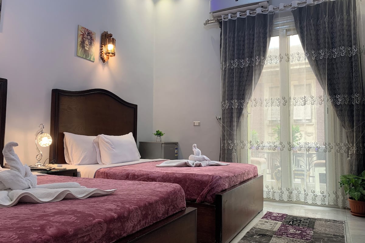 A hotel room with two beds, a nightstand with a lamp, a small table with a plant, and a window with curtains.