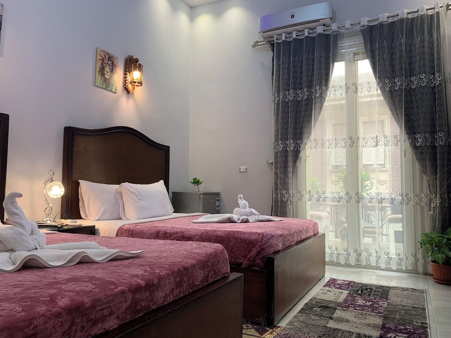 A hotel room with two beds, a nightstand with a lamp, a small table with a plant, and a window with curtains.