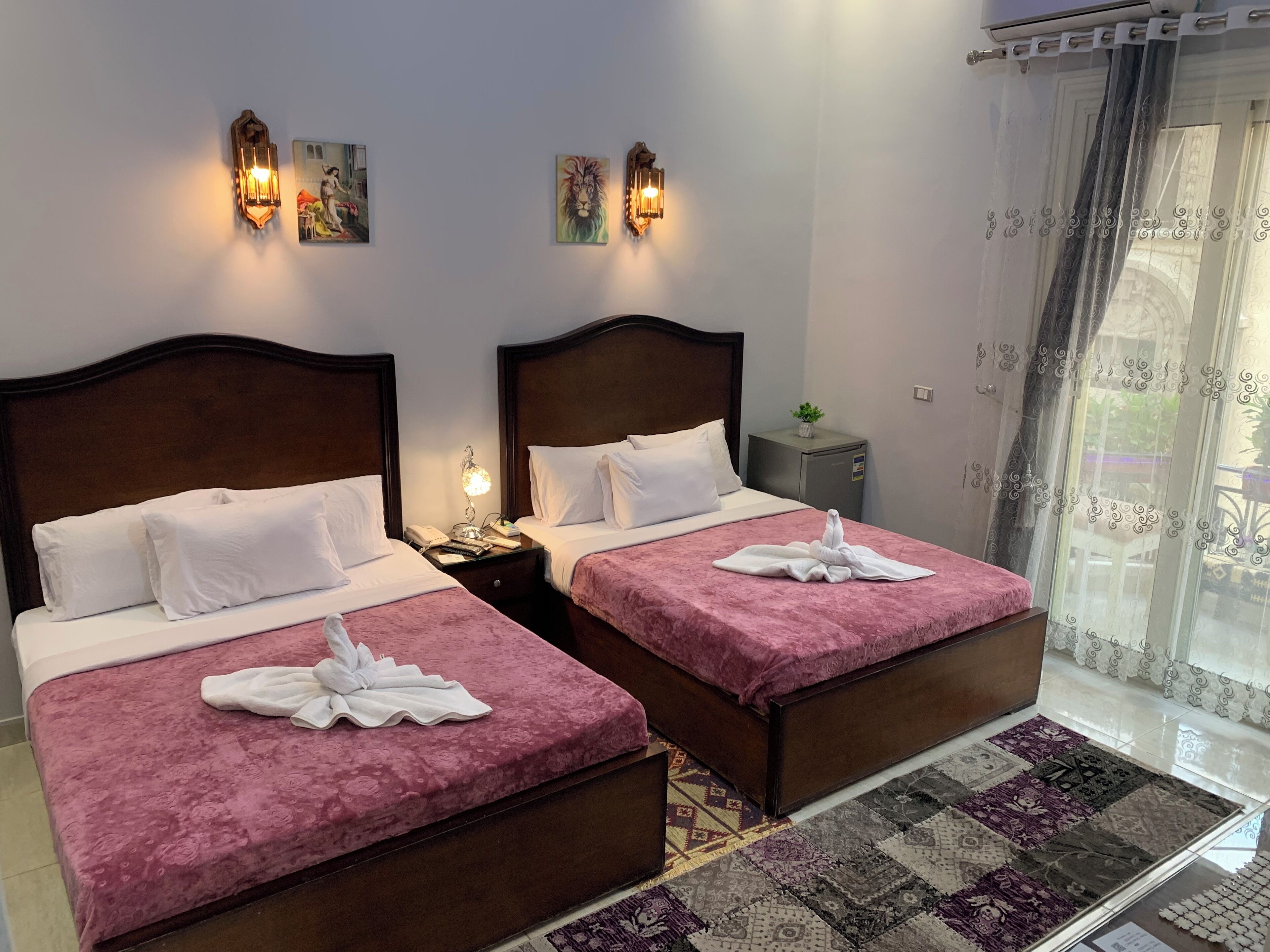 Habitación de hotel con dos camas, cada una con colcha rosa y almohadas blancas. Hay lámparas de pared y una pequeña mesa con una planta.