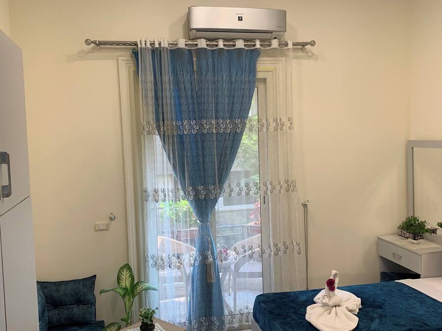 A hotel room with two beds, a small table with a plant, a blue curtain, and an air conditioning unit.