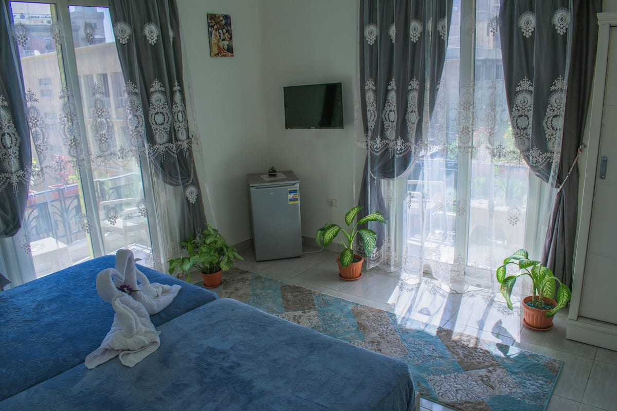 A hotel room with a bed, a TV, two potted plants, and a small refrigerator.