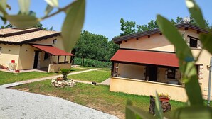 Un barrio residencial con dos casas, cada una con techo rojo y paredes color crema. Hay plantas en macetas y un pequeño jardín frente a las casas.
